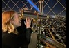 A woman standing on the summit of the Eiffel Tower looking through a telescope at night.