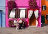Two women sitting on a bench in Burano in front of colorful houses.